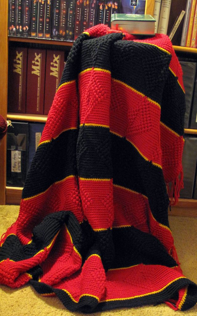 The blanket features wide alternating red and black stripes, each separated by thin yellow stripes. The red has a warm, rich tone, while the black create a striking contrast. The yellow stripes add a pop of color, giving the blanket a vibrant and dynamic look. It’s a cozy and visually interesting piece! The 3 dimensional designs are alternation triangles. 