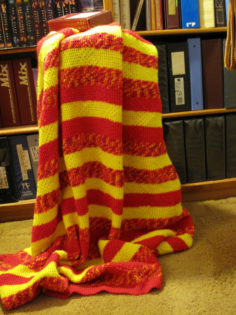 The blanket features alternating horizontal stripes of red and yellow, with some speckled patterns on the stripes. It looks both vibrant and cozy with alternating triangle and thatched patterns to touch. 