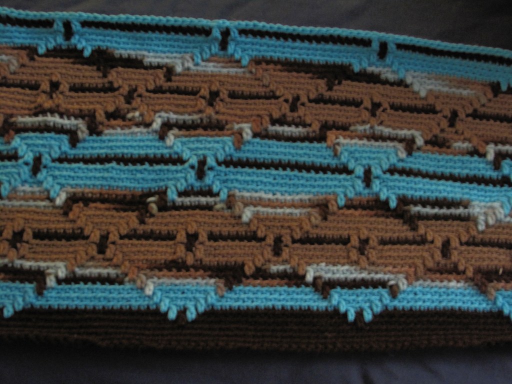 The blanket features a striking geometric pattern in shades of brown, blue, and light brown. Its interlocking shapes create a visually appealing design, making it both stylish and cozy. The texture looks thick and warm—perfect for colder weather. This blanket's craftsmanship and intricate design reflect the skill of its creator and the artistry involved in crocheting.