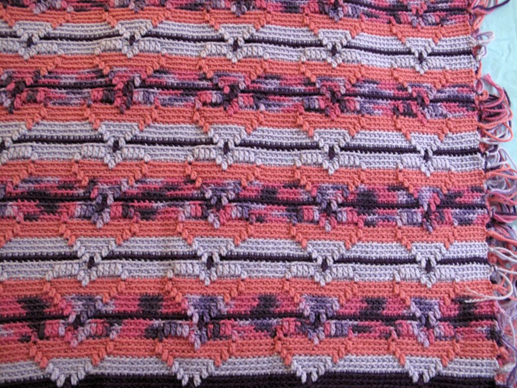 In the image, the blanket features an intricate geometric pattern. It has rows of zigzag shapes in pink, purple, and gray hues, creating a dynamic and appealing design.  The 3 dimensional pattern showcases stacked hearts. 