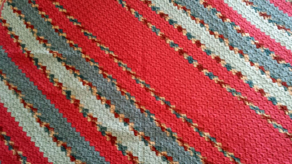 The image shows a close-up view of a tightly woven Afghan with a diagonal striped pattern. The stripes are composed of various colors, including salmon red, dark & light blue-gray, and some hints of tan. The pattern appears to be repetitive and geometric. The image is interesting due to the intricate design and the combination of colors, which create a visually appealing and dynamic pattern