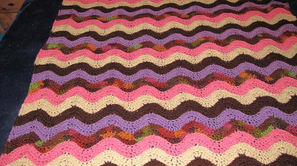 The blanket shows a ripple or wave pattern with alternating stripes of pink, purple, brown, beige, and multicolored yarn. The zigzag effect adds a lively touch and is visually engaging.