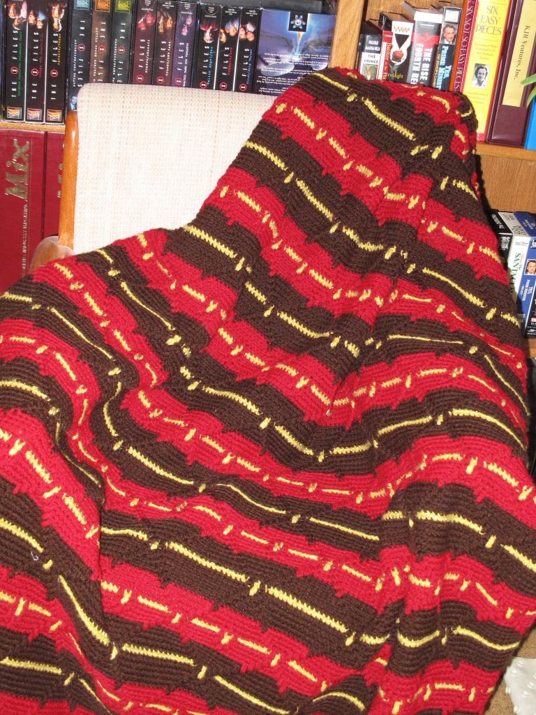The blanket in the image features alternating horizontal stripes of red, brown, and yellow. It's knitted or crocheted, with each stripe adding to its vibrant look. The combination of different yarns creates a soft and slightly bumpy surface, perfect for snuggling up on a cold day. It looks thick and warm, offering both visual and tactile comfort.
