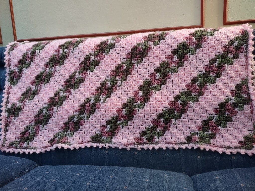 The texture of the blanket is distinctly crocheted, creating a grid-like stitch pattern. It’s detailed and slightly raised, adding a tactile quality to the diagonal stripes of pink, green, and maroon. The scalloped pink edges give it an additional layer of intricacy and softness, making it not just visually appealing, but also inviting to touch.