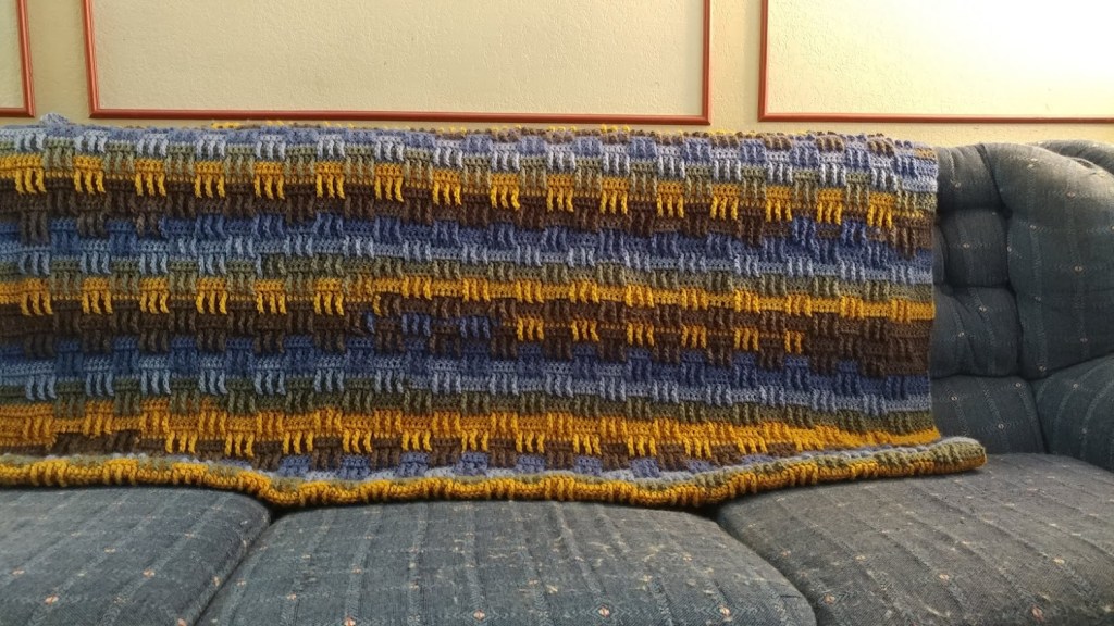 The blanket features a captivating horizontal striped pattern with colors such as blue, brown, green, and yellow. Each stripe appears to be made with a basket weave or similar stitch, giving it an intricate texture.